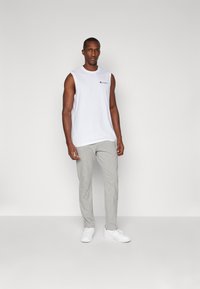 Witte mouwloze shirt met een logo, gecombineerd met lichtgrijze sweatpants en witte sneakers, staand tegen een egale achtergrond.
