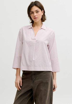 Žena s krátkými hnědými vlasy, oblečená v světle růžové pruhované blůze s dlouhým rukávem a tmavě hnědých kalhotech, stojící před jednobarevným pozadím.