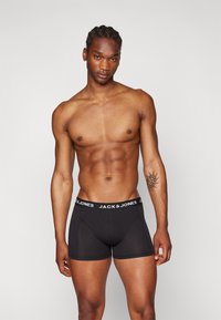 Boxeri negri cu un elastic alb inscripționat "JACK & JONES." Țesătura pare netedă, cu un design ajustat, evidențiind fizicul unui model masculin.