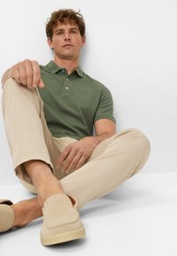 Green polo shirt with short sleeves, beige chinos, and light beige slip-on shoes. The shirt features a three-button placket and logo detail.