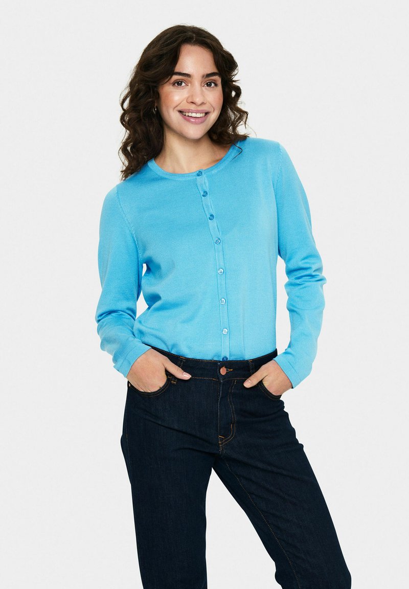 Vaaleansininen neuletakki, jossa on pyöreä kaula-aukko, napitus edessä ja pitkät hihat, yhdistettynä tummiin farkkuihin.