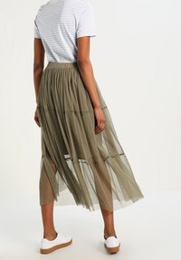 Falda larga de tul verde olivo con diseño en capas, cinturilla ajustada, textura transparente y longitud hasta la mitad de la pantorrilla, combinada con una camiseta a rayas y zapatillas.