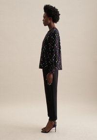 Top nero con motivo floreale colorato, vestibilità ampia, maniche lunghe; abbinato a pantaloni neri a gamba stretta e tacchi neri con cinturini.