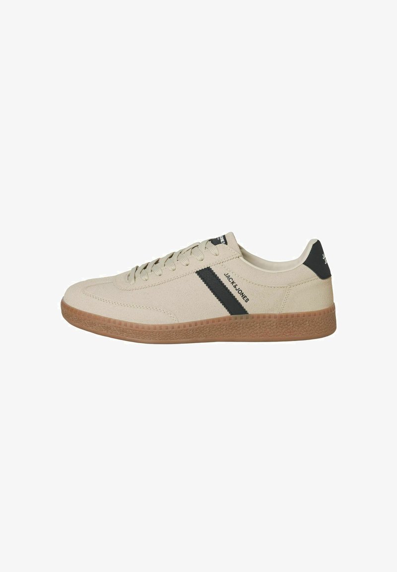 Beige sneakers with a textured canvas upper, contrasting black stripe, and rubber gum sole. Features classic lace-up design and branding detail.