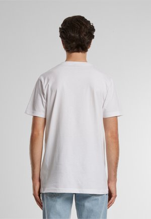 Homme aux cheveux bruns courts portant un t-shirt blanc uni à manches courtes et un jean bleu clair, vu de dos sur un fond gris.