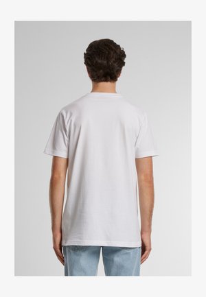 Mand med kort brunt hår iført en almindelig hvid T-shirt med korte ærmer og lyseblå jeans, set bagfra mod en grå baggrund.