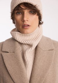 Beżowy wełniany płaszcz z dużymi klapami, nałożony na prążkowany golf w jaśniejszym odcieniu. Dziana czapka uzupełnia teksturowany wygląd.