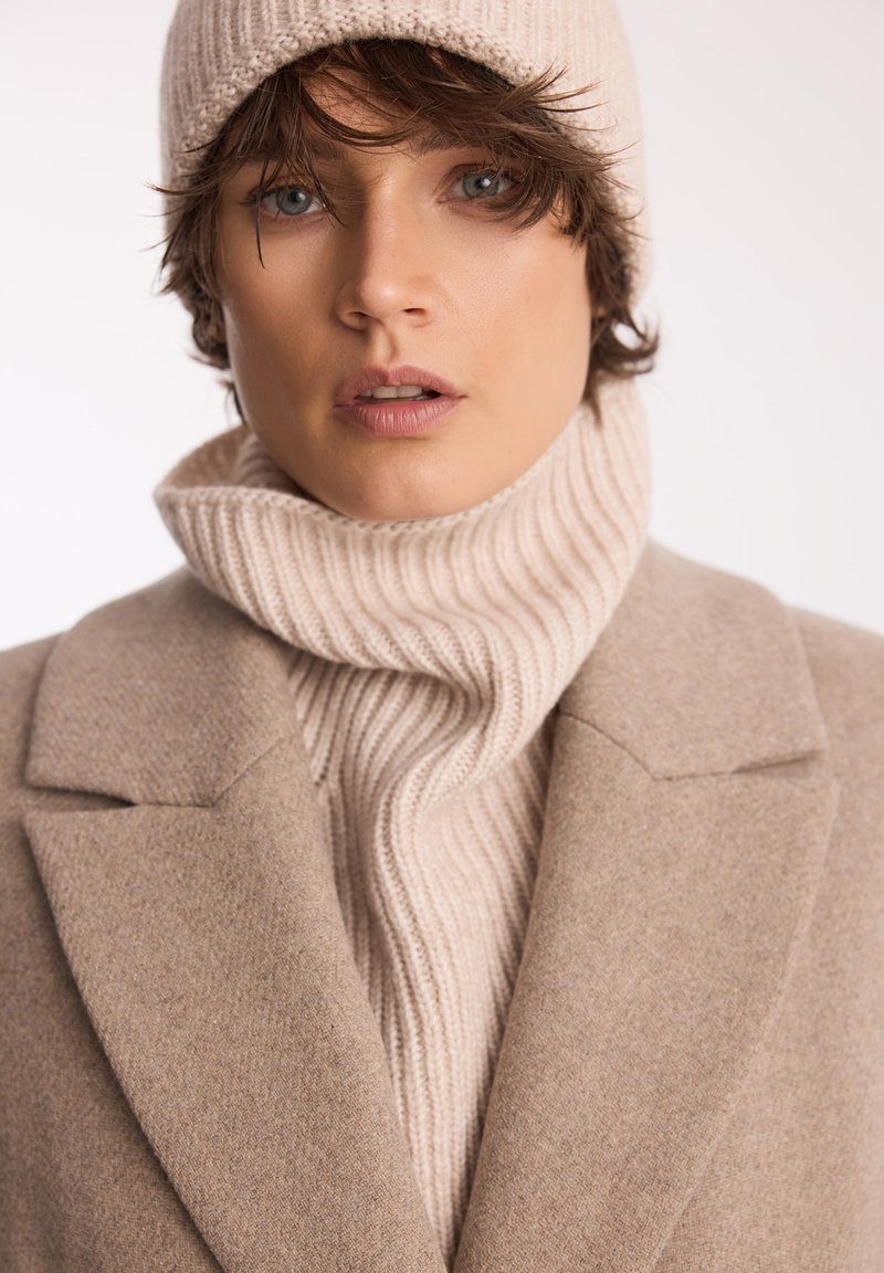 Beżowy wełniany płaszcz z dużymi klapami, nałożony na prążkowany golf w jaśniejszym odcieniu. Dziana czapka uzupełnia teksturowany wygląd.