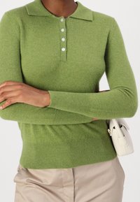 Osoba ubrana w zielony sweter polo z długimi rękawami i białymi guzikami, beżowe spodnie, trzymająca małą białą torebkę pod pachą.