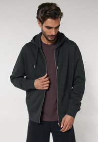 Sötétszürke, cipzáras kapucnis pulóver, állítható kapucnival, kenguru zsebekkel és bordázott mandzsettával, gesztenye színű póló fölött viselve. Simább anyagtextúra.