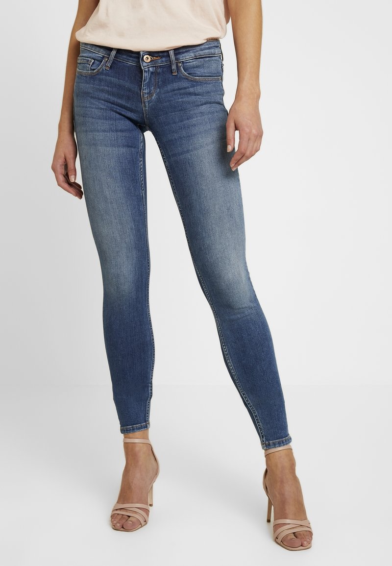 Jeans de denim azul ajustados con un acabado desgastado, cinco bolsillos, pasadores para cinturón y cremallera. Combinados con tacones nude de tiras.