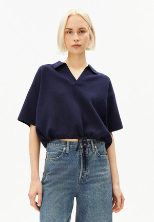 Jeune femme aux cheveux blonds courts portant un polo bleu marine à manches courtes noué à la taille et un jean bleu taille haute.