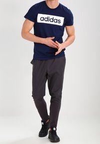 Mörkblå Adidas t-shirt med vit logotyp, parat med mörkgrå byxor och svarta sneakers. Modellen poserar med händerna på höfterna.