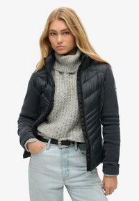 Chaqueta puffer negra superpuesta a un forro polar gris oscuro, con un alto cuello, cierre de cremallera y puños acanalados. Combinada con un suéter de punto gris claro.