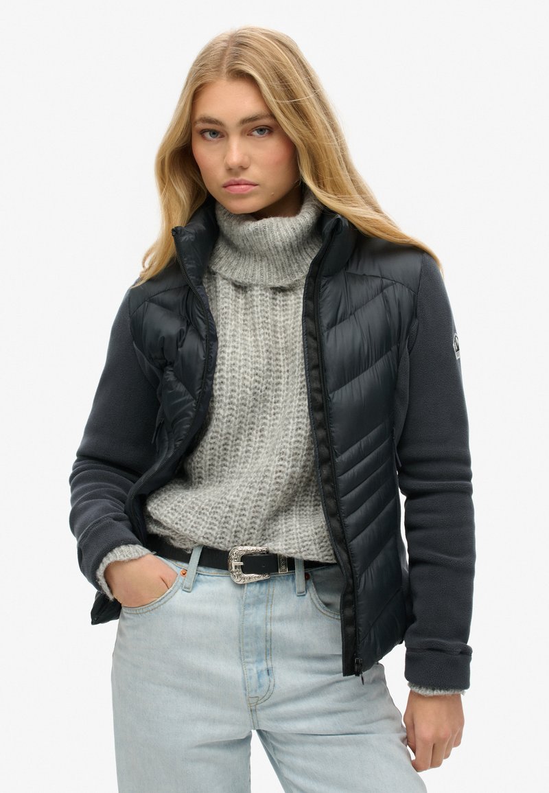 Chaqueta puffer negra superpuesta a un forro polar gris oscuro, con un alto cuello, cierre de cremallera y puños acanalados. Combinada con un suéter de punto gris claro.