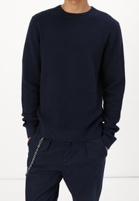 Námořnicky modrý pletený svetr s kulatým výstřihem, žebrovanými manžetami a texturovaným povrchem, doplněný tmavými kalhotami s akcentem stříbrného řetízku.