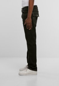 Jeans de denim negro con detalles de costura blanca, diseño de pierna recta, cintura a media altura, usados con zapatillas blancas. Vista de perfil lateral.
