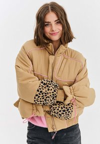 Mladá žena sa usmieva, má krátke hnedé vlasy a na sebe má hnedý puffesový kabát s ružovým lemovaním a rukávmi so leopardím vzorom, ktorý nosí cez ružový sveter.