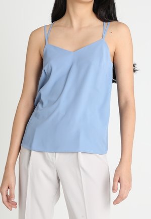 Femme portant un caraco bleu clair sans manches avec double bretelles fines et un pantalon blanc taille haute sur fond uni.