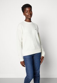 White knitted sweater with ribbed cuffs and hem, featuring a small green logo on the chest, paired with dark blue jeans.