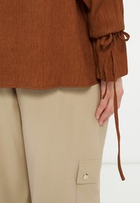 Καφέ υφασμάτινη μπλούζα με μακριά μανίκια και δετούς μανσέτες, συνδυασμένη με μπεζ παντελόνι που διαθέτει πλαϊνή τσέπη με κούμπωμα κουμπί πατιλέτα.