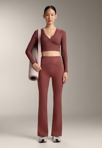 Donna in piedi con un tappetino da yoga arrotolato sulla spalla, indossa un crop top a maniche lunghe color ruggine e pantaloni svasati abbinati con scarpe da ginnastica.