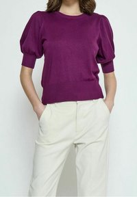 Lila gestrickter Pullover mit kurzen Puffärmeln sowie Rippkragen und -saum. Kombiniert mit cremefarbenen Hosen, Hände in den Taschen.