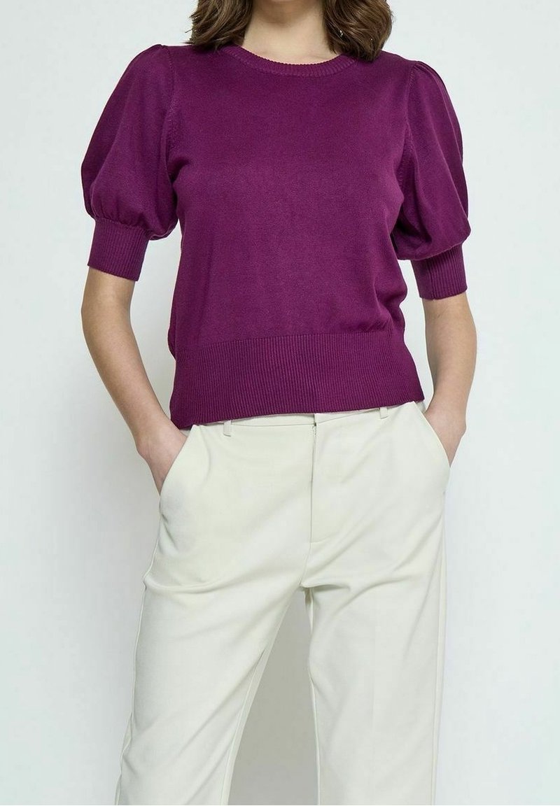 Lila gestrickter Pullover mit kurzen Puffärmeln sowie Rippkragen und -saum. Kombiniert mit cremefarbenen Hosen, Hände in den Taschen.