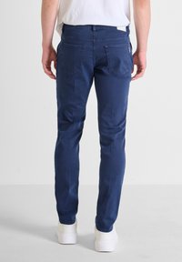 Tmavě modré džíny s úzkým střihem, vybavené dvěma zadními kapsami, decentním prošíváním a hladkou texturou. Nosené s bílými botami.