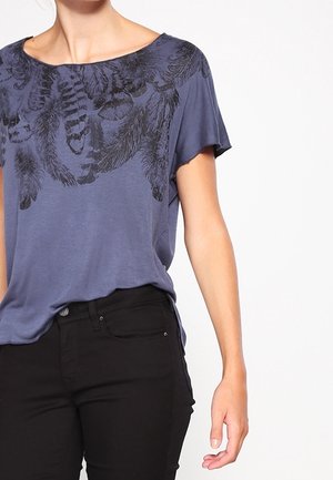 Marineblauwe t-shirt met korte mouwen, voorzien van een zwart verenpatroon over de borst. Gecombineerd met zwarte nauwsluitende broek, met een gladde textuur.