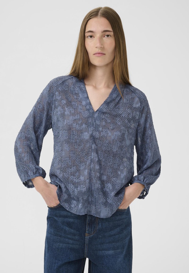 Světle modrá blouse s potiskem, výstřihem do V, dlouhými rukávy a nařasenými manžetami. Vyrobena z průhledného materiálu s jemnou strukturou.