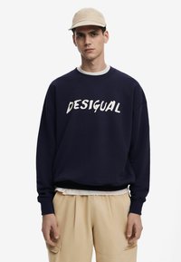Sudadera azul marino con el texto "DESIGUAL" en blanco, cuello redondo, puños de canalé, superpuesta sobre una camisa blanca, combinada con pantalones beige.