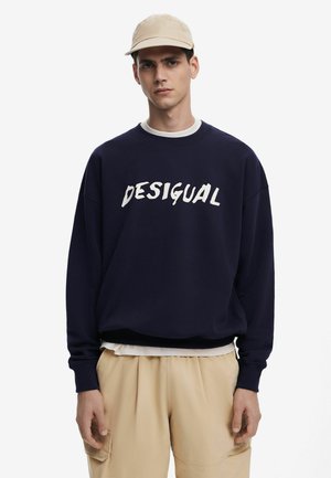 Marineblauwe sweatshirt met witte "DESIGUAL" tekst, ronde hals, geribbelde boorden, over een wit shirt gedragen, gecombineerd met beige broeken.