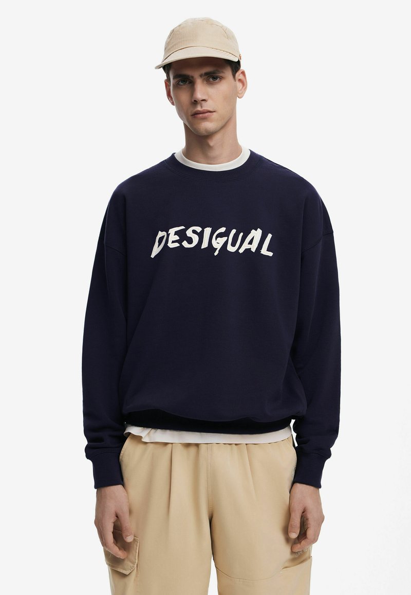 Sudadera azul marino con el texto "DESIGUAL" en blanco, cuello redondo, puños de canalé, superpuesta sobre una camisa blanca, combinada con pantalones beige.