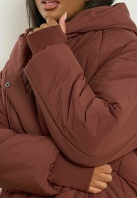 Brązowa pikowana kurtka z kapturem, prążkowanymi mankietami i guzikami. Materiał jest gładki z lekkim połyskiem i puszystą strukturą.