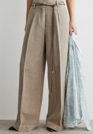 Beige bukser med brede ben og et tyndt brunt bælte i taljen, kombineret med mørke sko, der holder et lyseblåt mønstret stof i højre hånd.