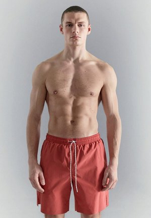 Giovane uomo muscoloso in piedi contro un muro grigio chiaro, indossa pantaloncini da bagno rossi con cordino bianco, volto verso l'obiettivo con espressione neutra.