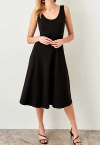 Femme portant une robe midi noire sans manches et des sandales noires à talons hauts avec des brides, debout contre un mur blanc.