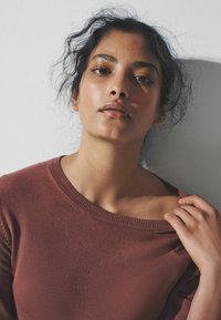 Kaštanový pletený svetr se širokým výstřihem, žebrovanými detaily na okrajích a měkkou texturou; modelka jemně upravuje výstřih jednou rukou.