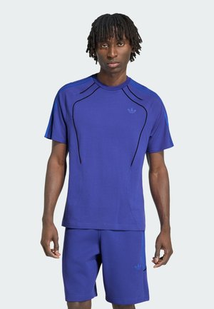 Junger Mann trägt ein blaues Sportshirt und passende Shorts mit schwarzen und blauen Streifendetails, steht vor einem schlichten hellen Hintergrund.