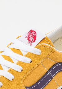 Žluté plátěné tenisky s bílými tkaničkami, které mají fialový boční pruh a bílou jazykovou etiketu s červeným logem Vans.