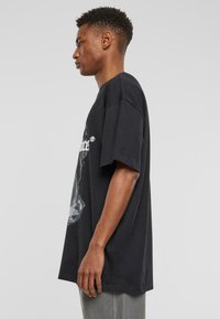 Melnas kokvilnas T-krekls ar īsām piedurknēm, brīvu piegriezumu un grafisku attēlu priekšpusē, attēlots sānos.