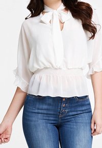 Weißes Bluse mit einer Schleifenbindung am Hals, halb durchsichtiger Stoff, geraffte Ärmel und eine elastische Taille, kombiniert mit dunkelblauen, eng anliegenden Jeans.