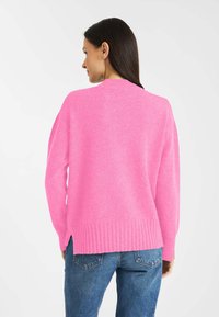 Różowy sweter z prążkowanym wykończeniem i mankietami, o luźnym fasonie oraz rozcięciami po bokach. Noszony z niebieskimi dżinsami, widok z tyłu.