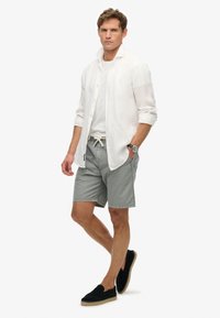 Chemise blanche à manches longues portée sur un t-shirt blanc, associée à un short gris et des espadrilles noires, avec un léger cordon de serrage et des poches latérales.