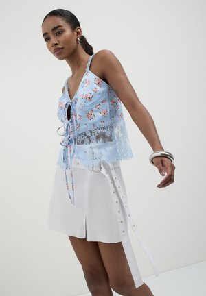 Donna che indossa un top in pizzo floreale blu e una gonna bianca con fibbia e cintura ad occhiello, in posa su uno sfondo grigio uniforme.