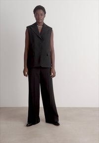 Gilet nero su misura con bottoni e rever, abbinato a pantaloni neri a gamba larga. Tessuto liscio e silhouette strutturata. Design minimalista.