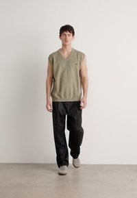 Top de malha sem mangas em verde oliva com decote em V e padrão texturizado, combinado com calças pretas e sapatos cinza claro, num fundo simples.