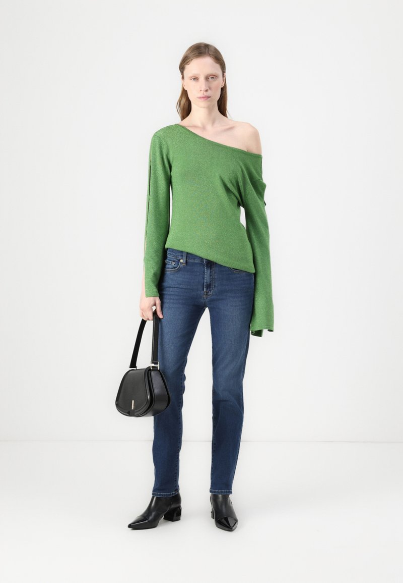 Grüner schulterfreier Strickpullover, schlanke blaue Jeans, schwarze Handtasche und spitze schwarze High Heels in einem minimalistischen Umfeld.
