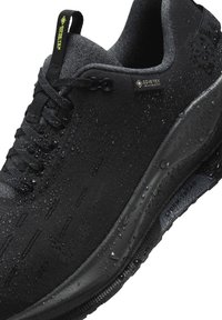 Chaussure de running noire étanche avec étiquette Gore-Tex, lacets noirs et gouttes d'eau sur la surface et la semelle.